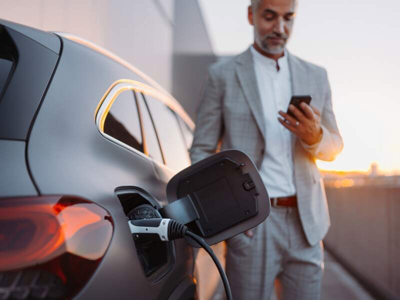 Grey-haired man in a grey suit jacket and trousers charging his electric vehicle at a shopping complex car park against a sunset backdrop.